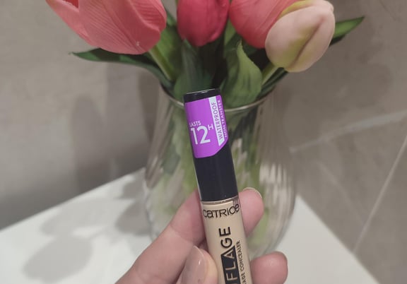 A hand holding a tube of Catrice Liquid Camouflage High Coverage Concealer in front of a vase of tulips. review