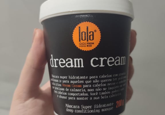 A hand holds a black tub of Lola Dream Cream deep conditioning hair masque with a white lid and orange logo. review